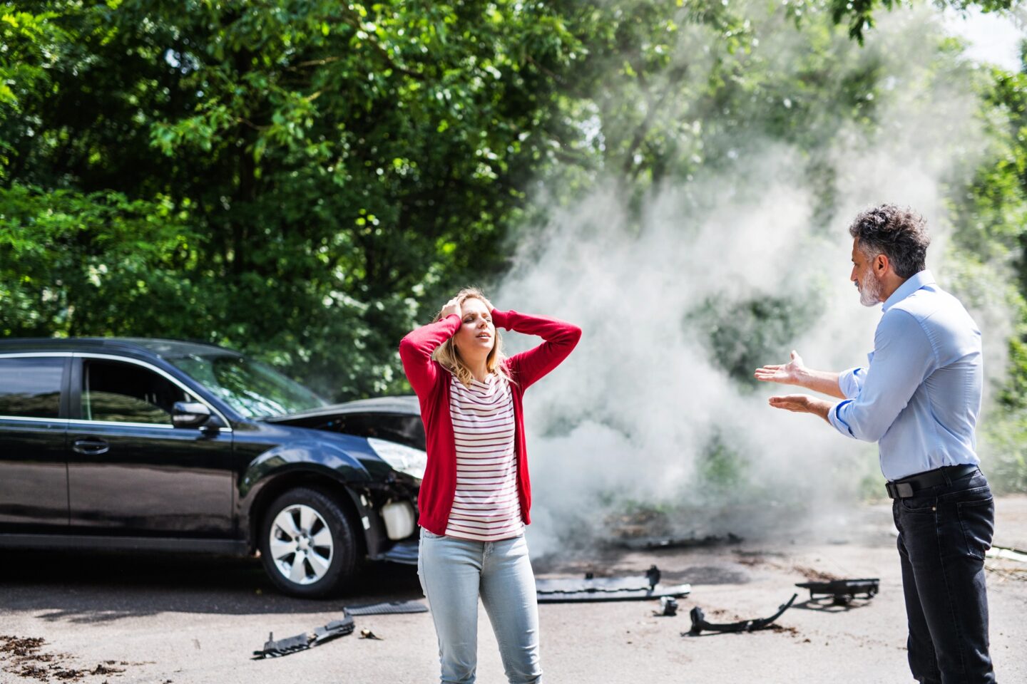 A man and woman standing in front of a smoking wrecked car on a road.