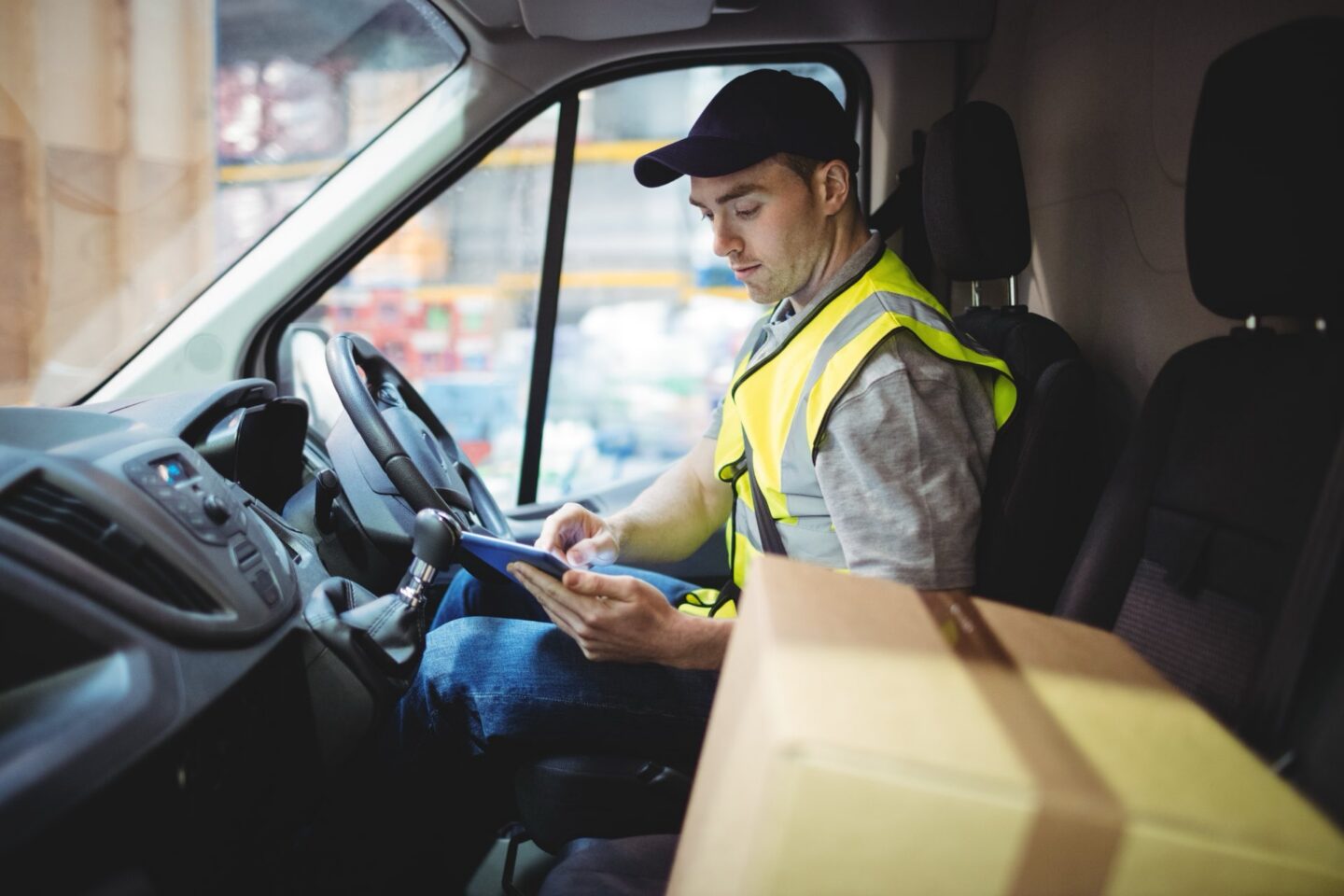 A delivery driver in a yellow vest holding a clipboard inside of his vehicle with a box next to him