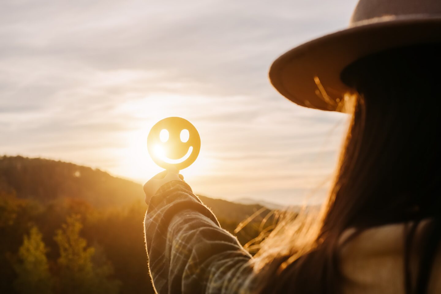 A woman holds a cardboard cutout of a smiley face up against the horizon with the sun behind it.