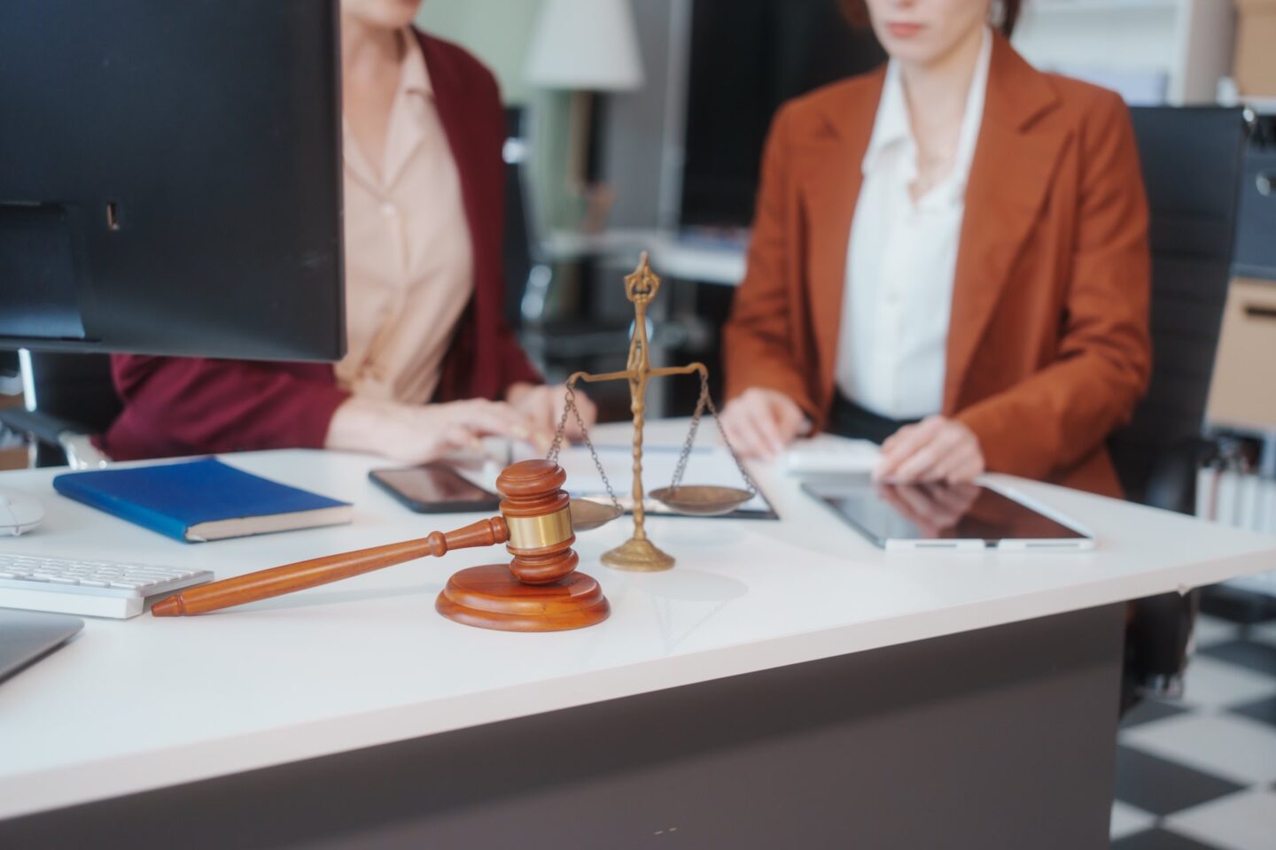 Two women meeting at a table in front of a computer next to a gavel and justice of scales
