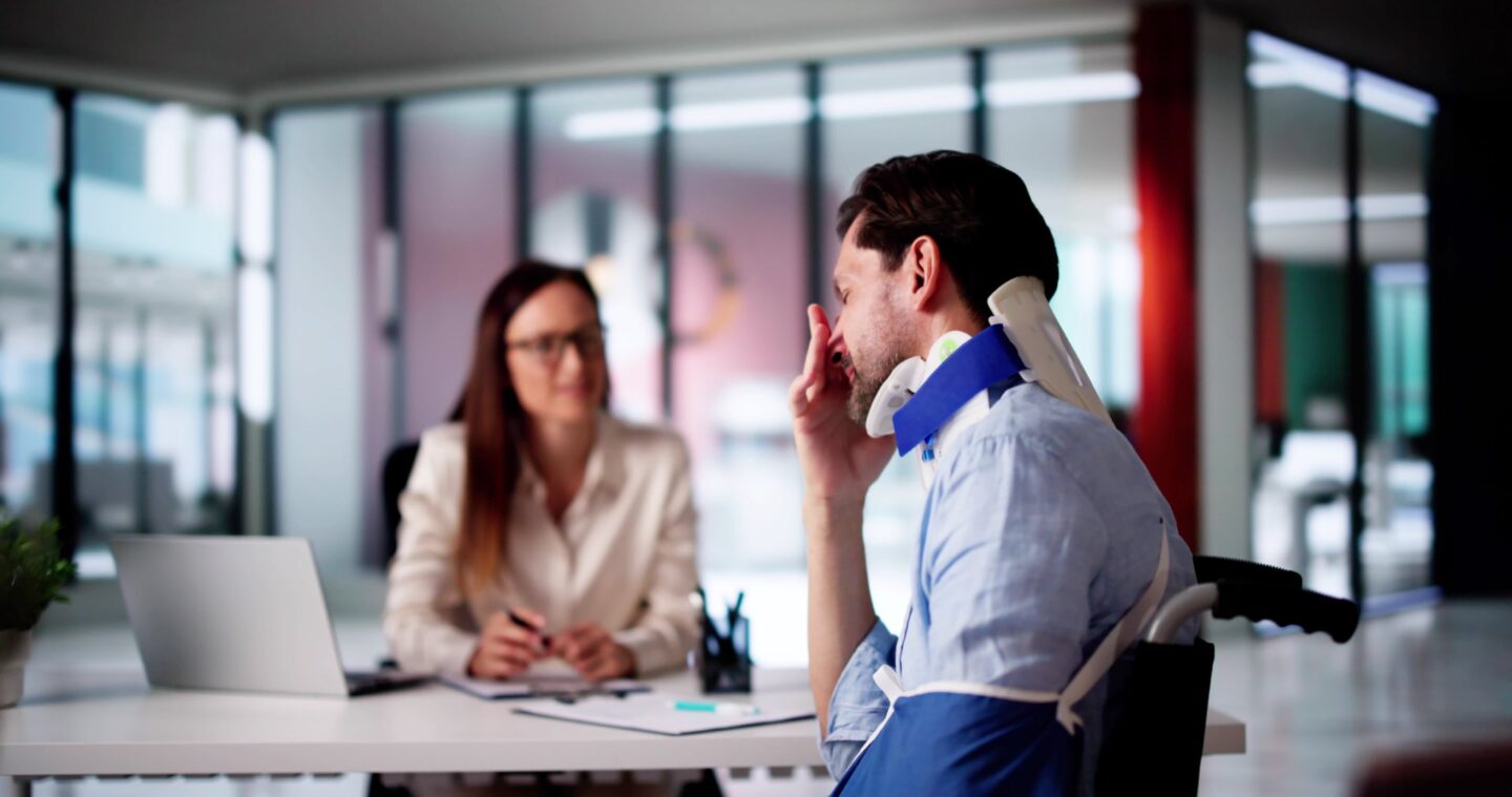 Woman holding a pen next to a laptop speaking with someone with a neck brace and arm sling on
