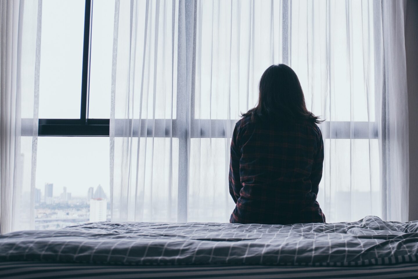 The silhouette of a woman sitting on a bed with her back facing the camera looking out a window in the dark.