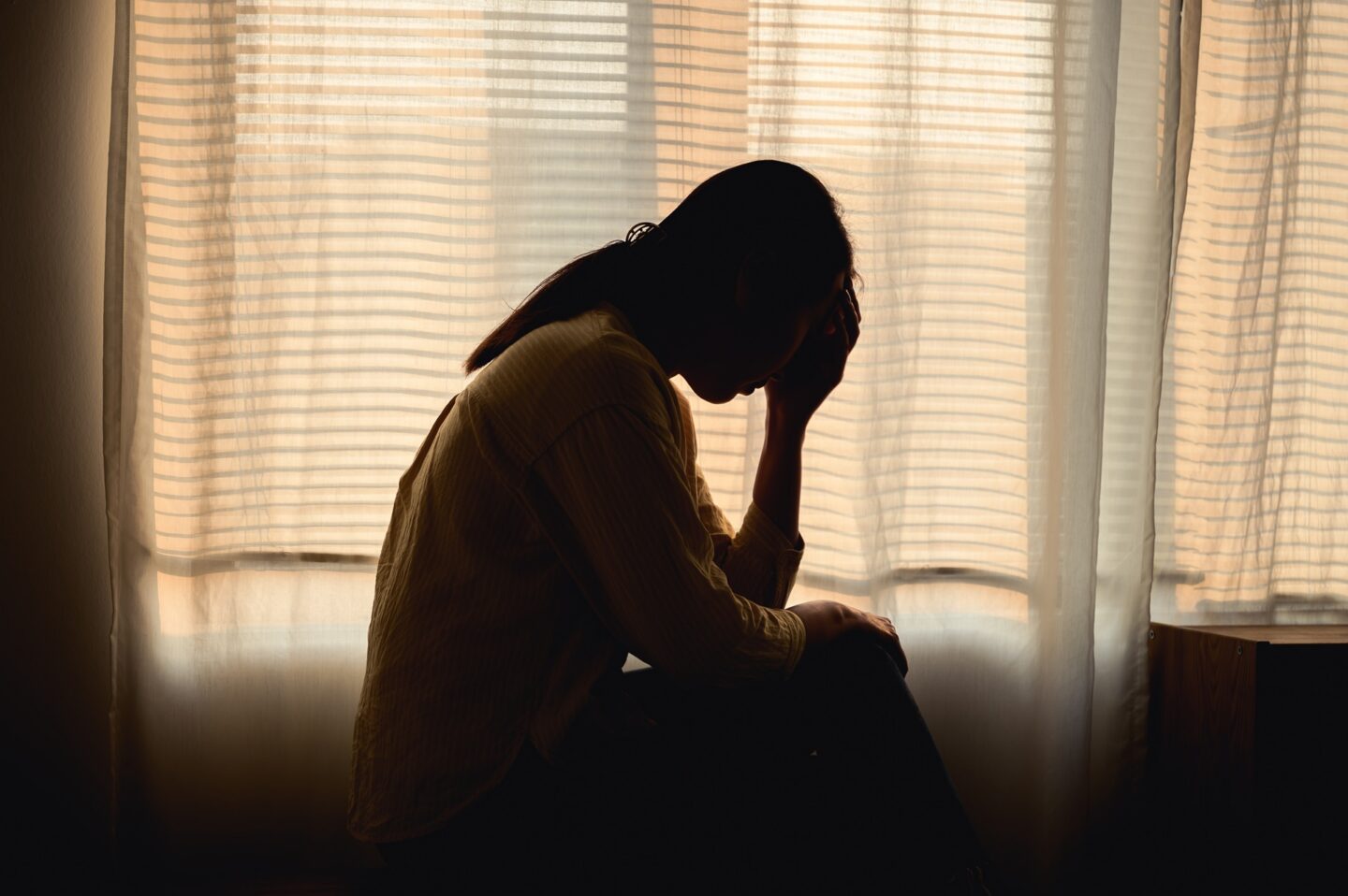 The silhouette of a woman sitting on a bed in a room with the curtains closed.