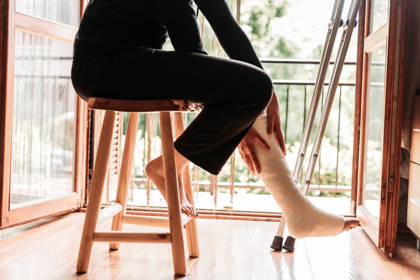 A woman in a leg cast sits on a stool next to crutches reaching down at her leg.